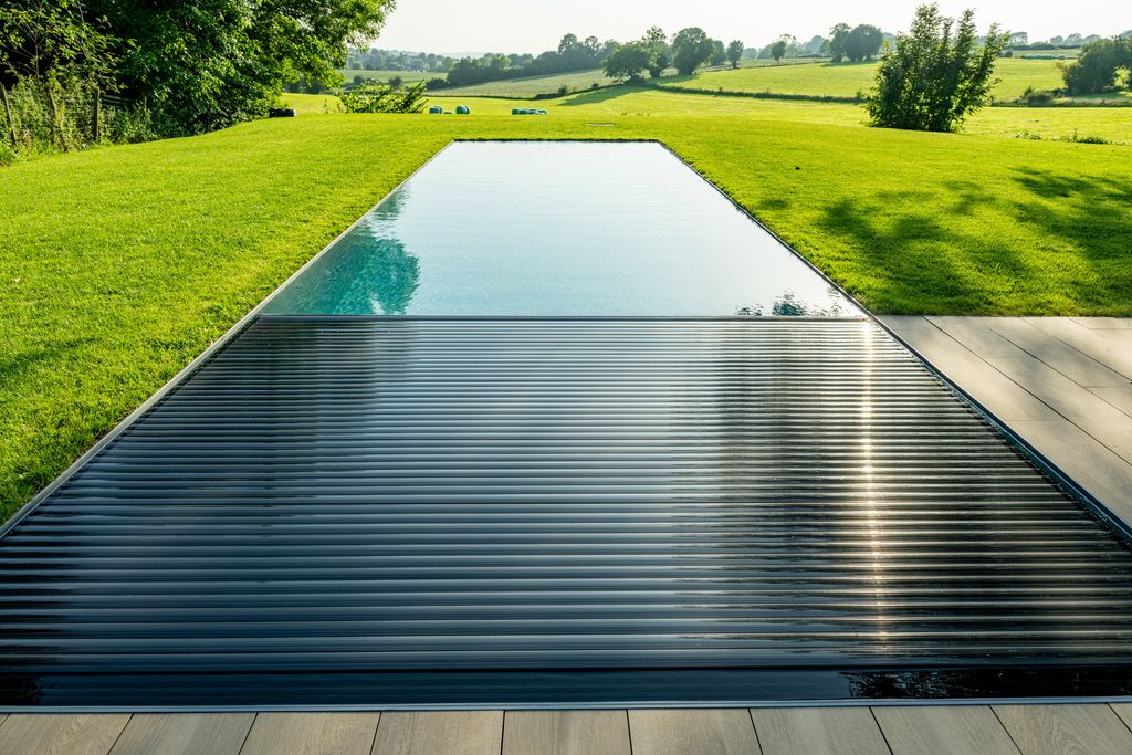 Le volet immergé partiellement déployé sur la piscine, vu depuis la terrasse, avec la pelouse et la campagne au-delà