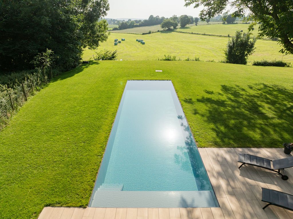 Vue aérienne d'une piscine à débordement intégrée au gazon, sans margelle, au milieu d'une grande pelouse face à la campagne de Verviers