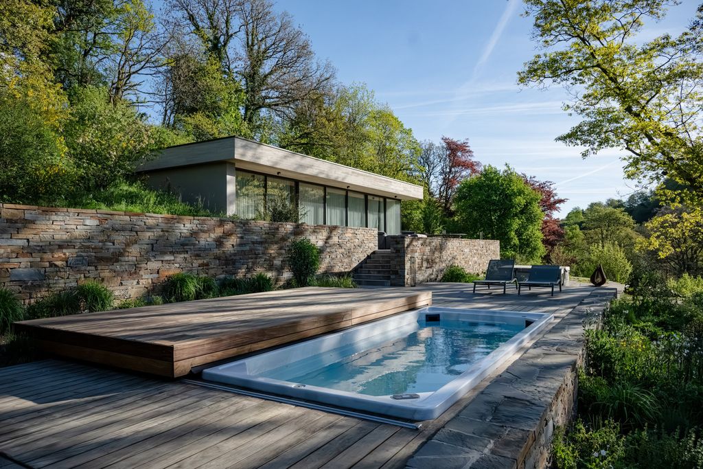 Piscine à volet en bois et pavillon vitré à toit végétalisé, adossés à un mur en pierre naturelle dans une colline boisée près d'Eupen