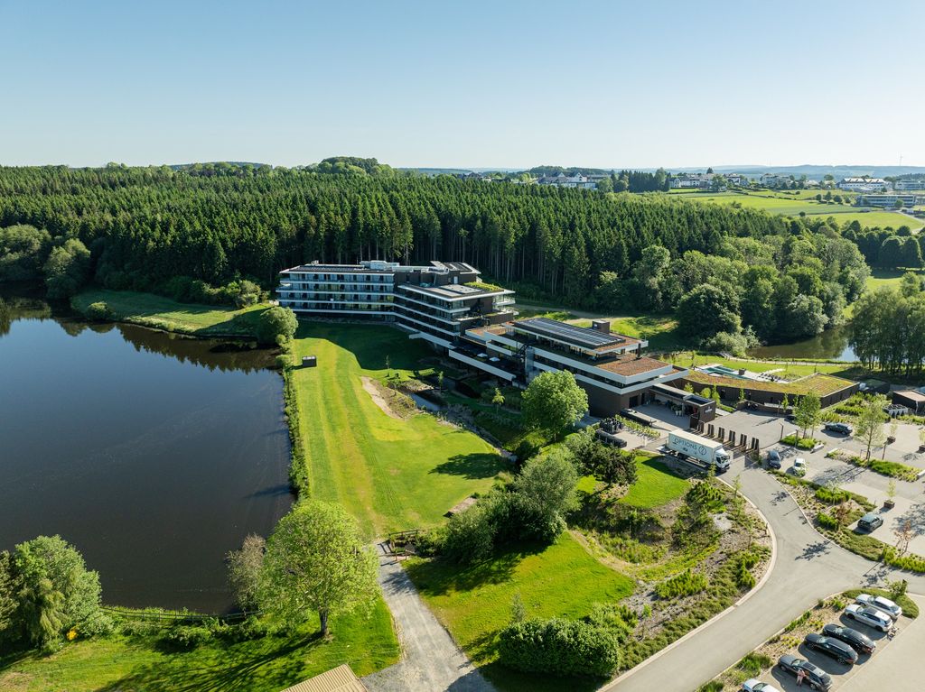 Vue aérienne de l'Anatura Hotel & SPA entre lac, pelouses et massifs plantés à Weiswampach